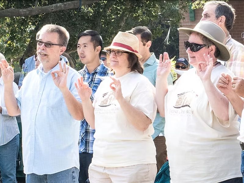 A group of people outdoors, wearing casual clothes and making hand gestures, with trees and buildings in the background on a sunny day.
