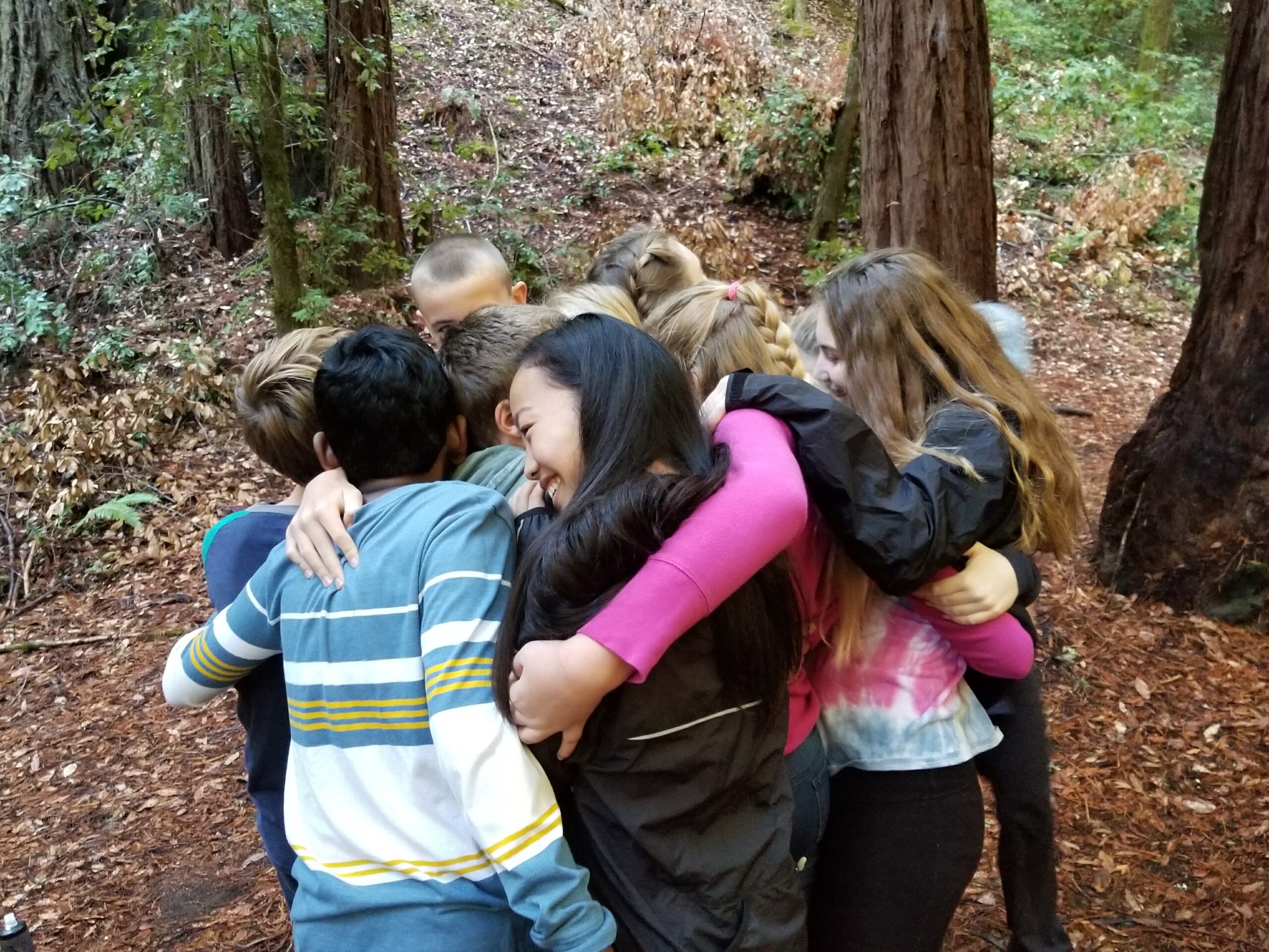 A group of young people embraces in a forest clearing, surrounded by tall trees and leafy undergrowth, fostering a sense of unity.