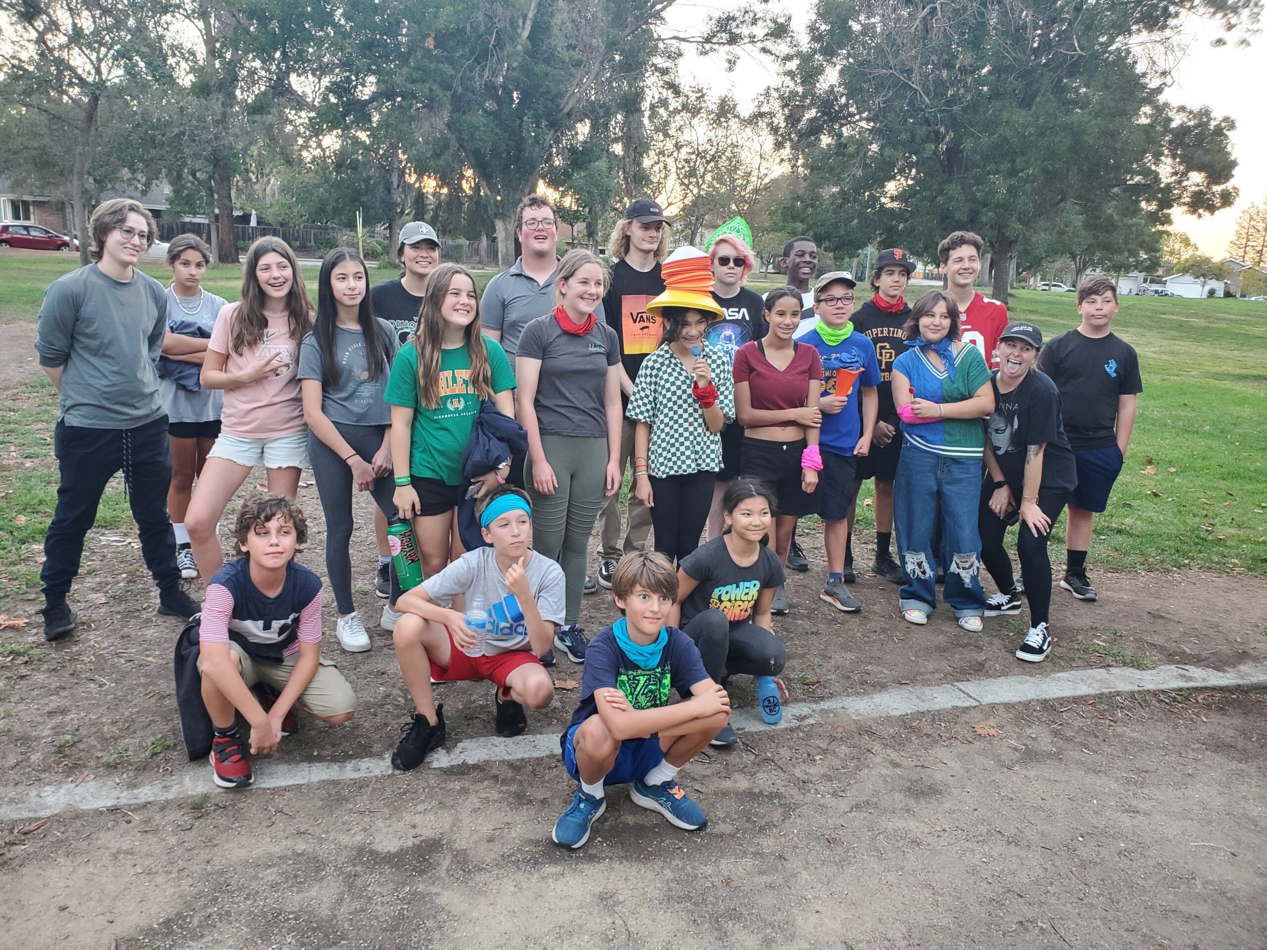 A group of people and children pose outdoors in a park, surrounded by trees, enjoying a social gathering under a serene sky.