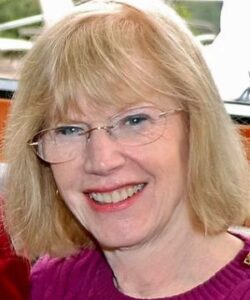 A smiling person with glasses and blonde hair is indoors. The background is blurred, showing some greenery. No landmarks or historical buildings visible.