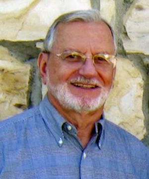 An older person with glasses, wearing a blue shirt, stands in front of a textured stone wall, smiling at the camera.