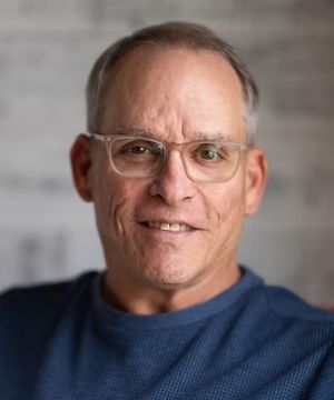 A person wearing glasses and a blue shirt smiles at the camera. The background is softly blurred, highlighting the person's face.