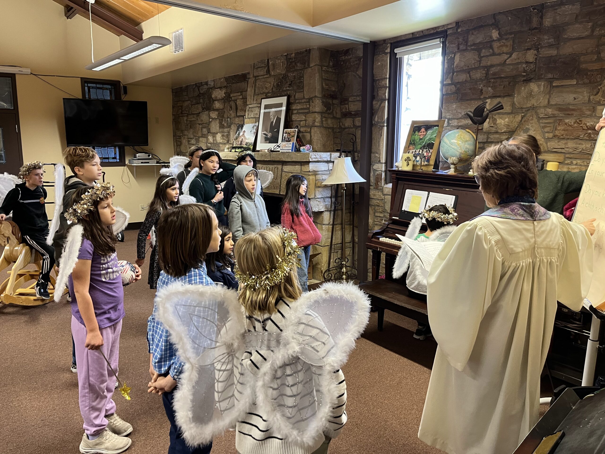 Children in angel costumes listen to a person in a robe playing piano in a room with stone walls and a TV.