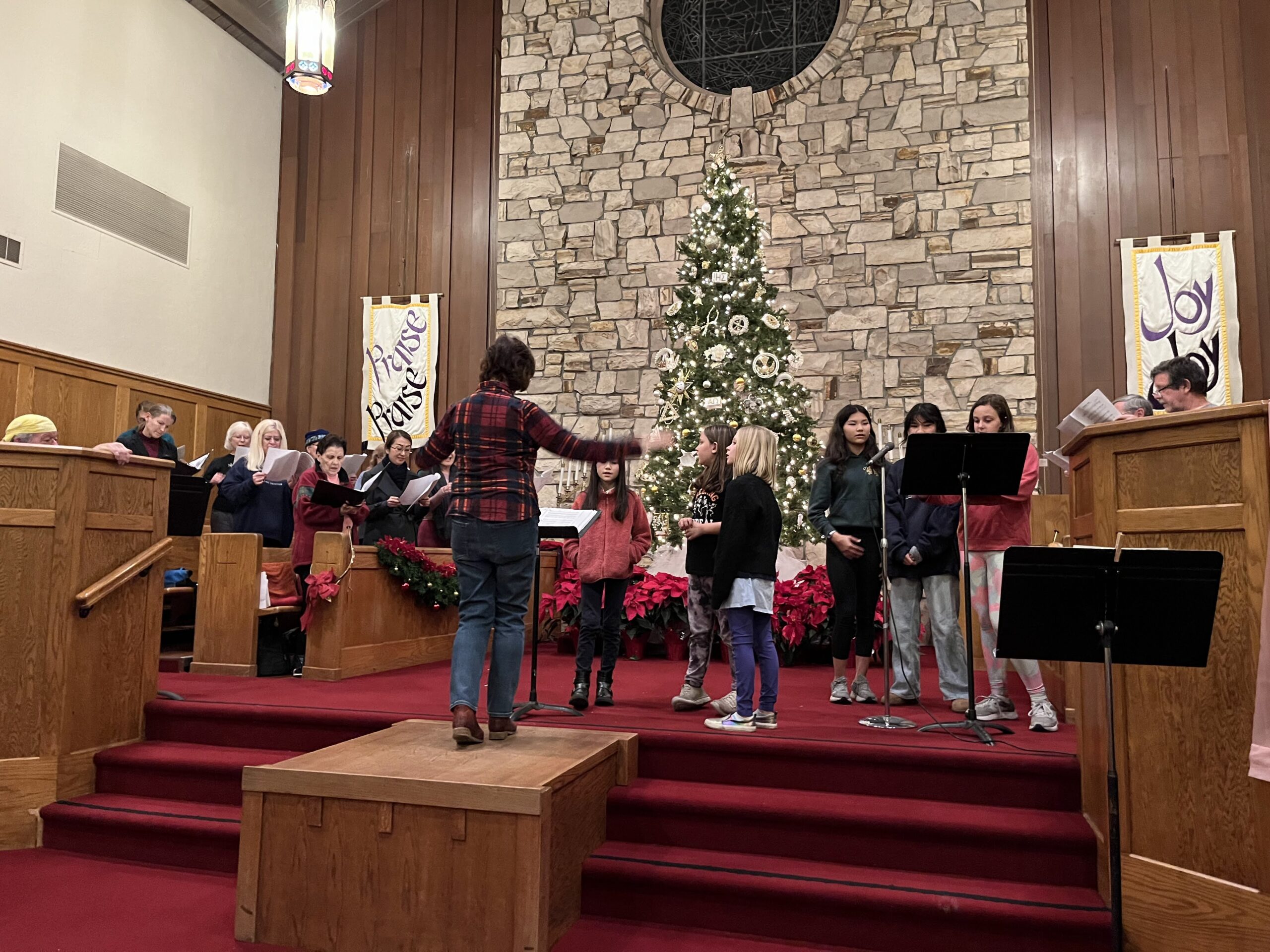 A choral group rehearses in a church adorned with Christmas decorations, featuring a lit Christmas tree and poinsettias.