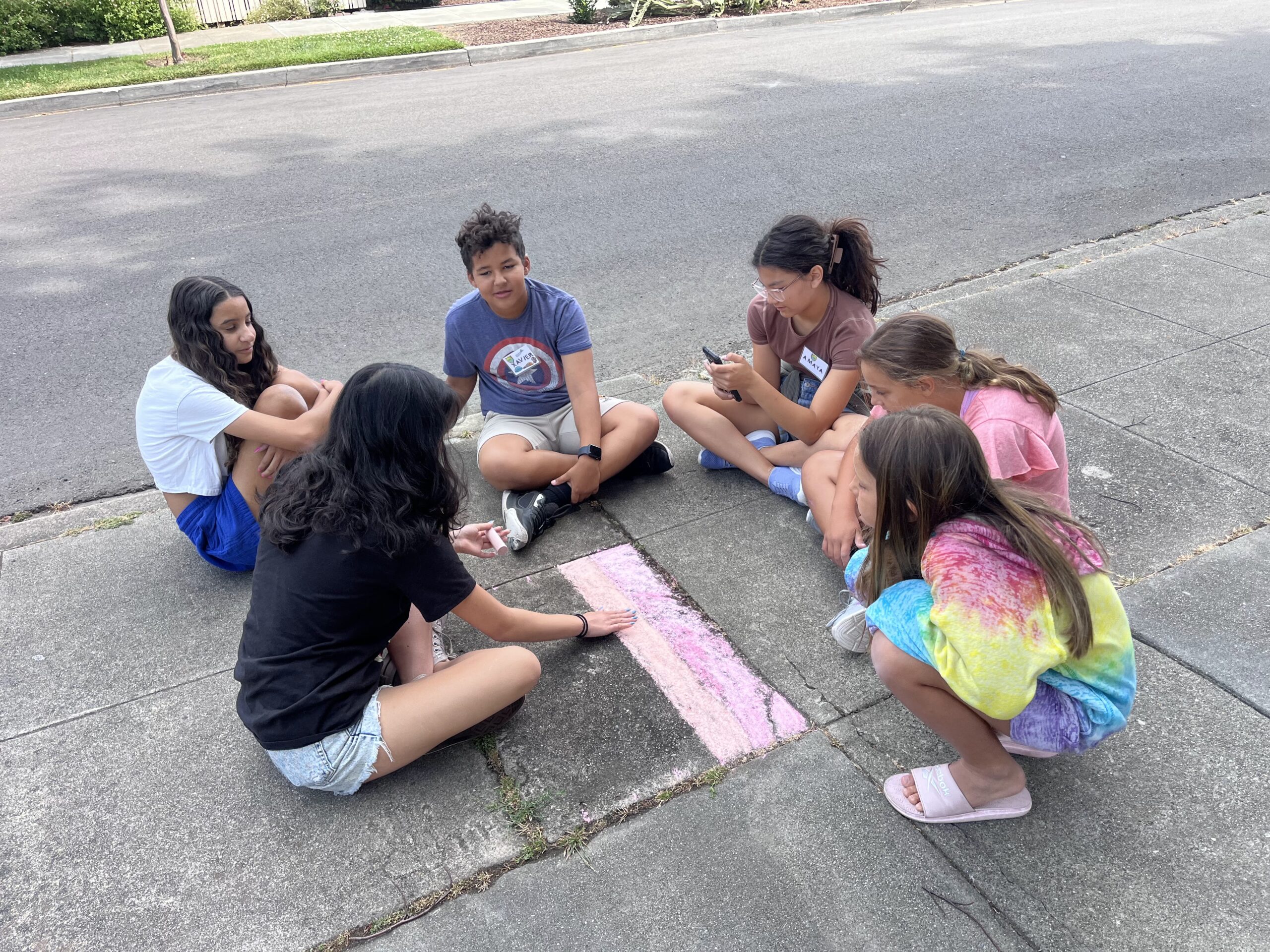 A group of children sit on the sidewalk, drawing with pink chalk, in a suburban neighborhood on a clear day.