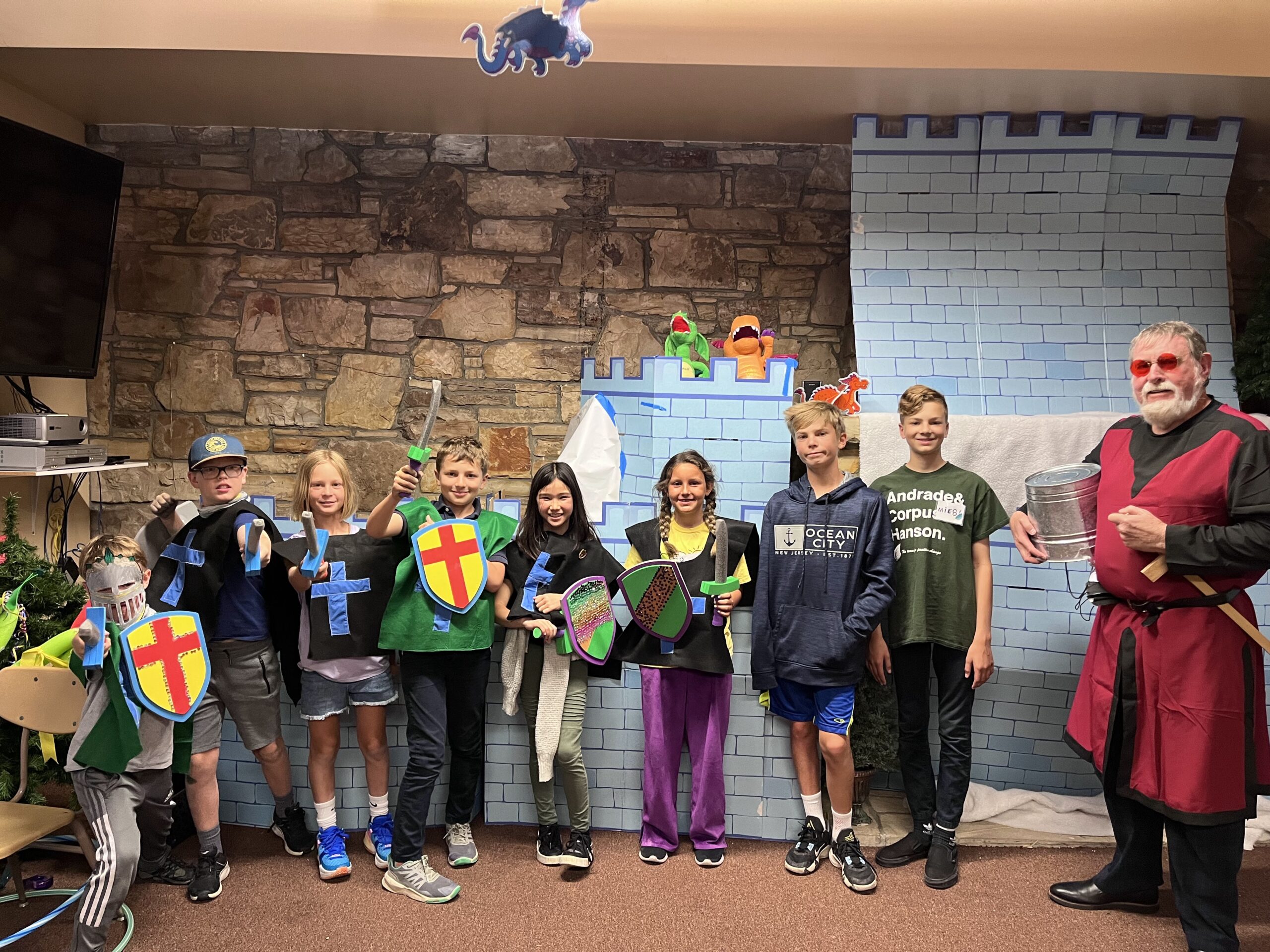A group of children, dressed as knights with shields and swords, pose in front of a castle-themed backdrop with a person nearby.