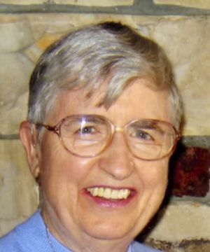 A person with short gray hair and glasses smiles in front of a stone wall. No landmarks or historical buildings are visible.