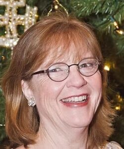 A person with glasses and earrings smiles in front of a decorated Christmas tree adorned with lights and ornaments.