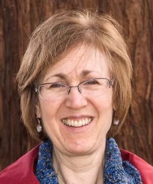 A person with short hair and glasses smiling, wearing a blue scarf and red jacket, set against a wooden background.