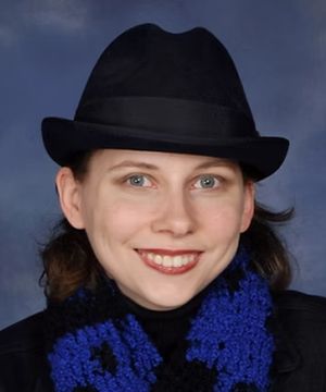 A person with a hat smiles at the camera against a blue background, wearing a black and blue scarf and jacket.