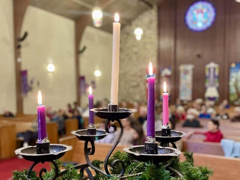 Five lit candles on an Advent wreath inside a church with stained glass windows, surrounded by people seated on wooden pews.