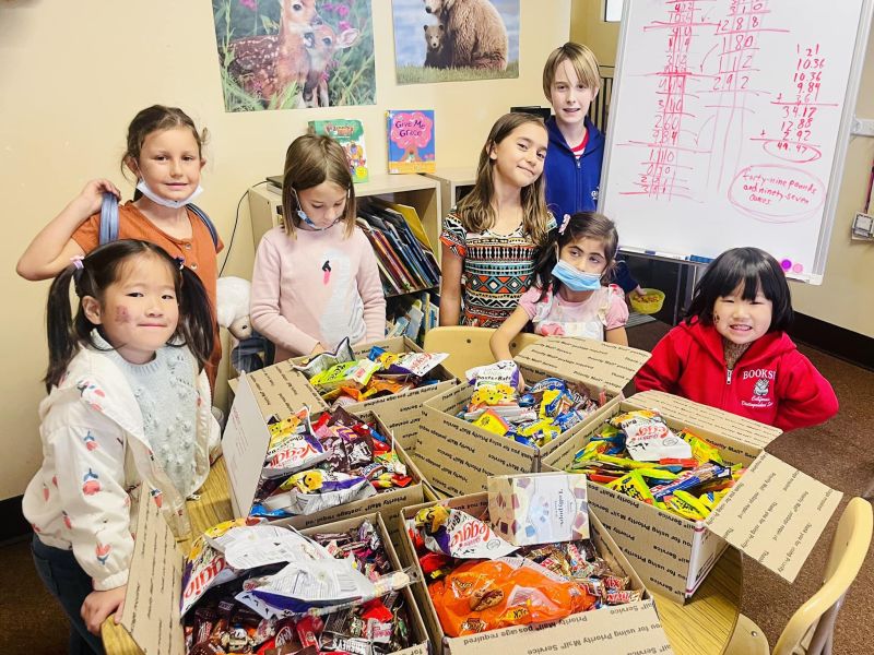 Children smiling around tables with open boxes filled with candy. Classroom setting with animal posters and whiteboard in the background.
