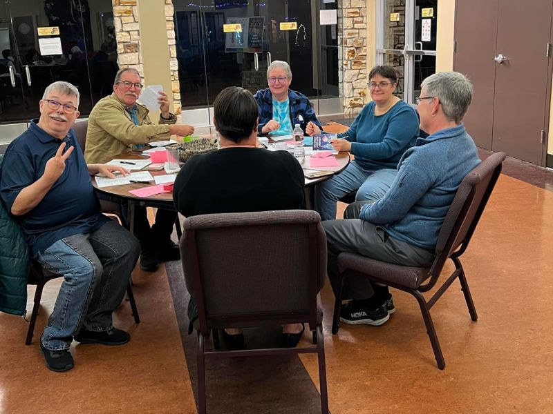 A group of six people sit around a table, talking and smiling, with papers and drinks in a casual indoor setting.