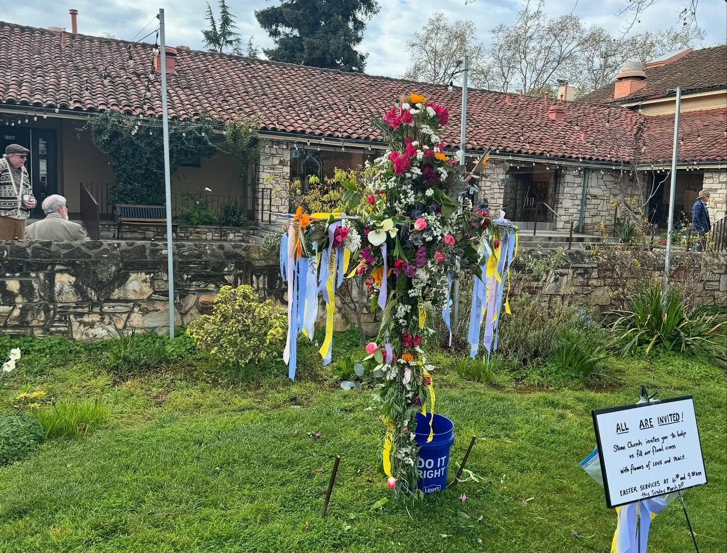 A decorated floral cross stands on grass, surrounded by a historic stone building. Two people converse nearby. A sign invites public participation.