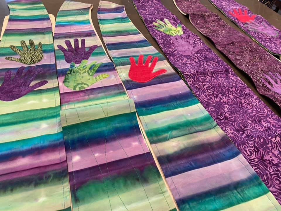 Colorful fabric strips with handprints are laid out in rows. The patterns and hues include purple, green, and red on a table.