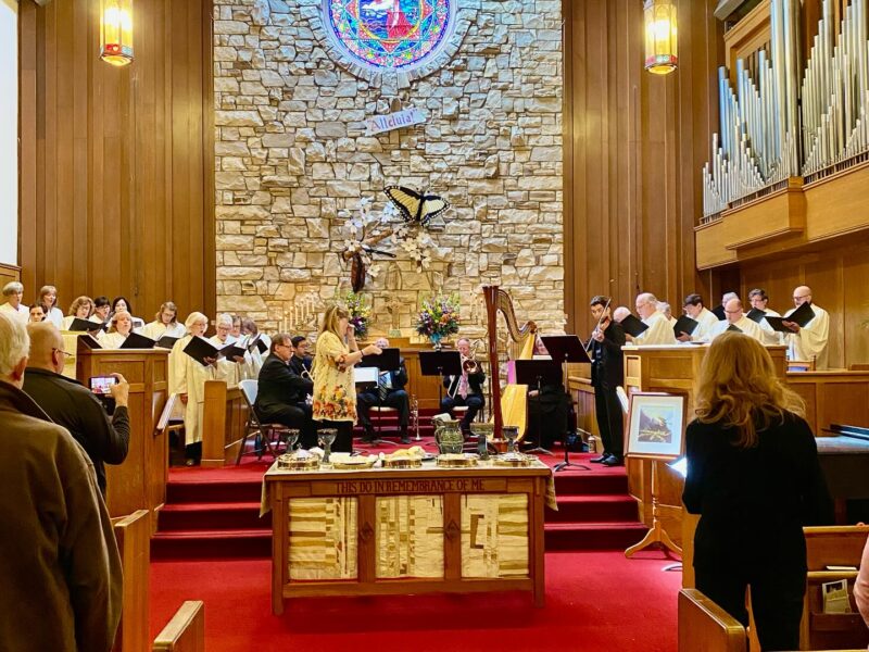 People singing in a church choir, surrounded by musical instruments and congregation. A stained glass window and stone wall are in the background.