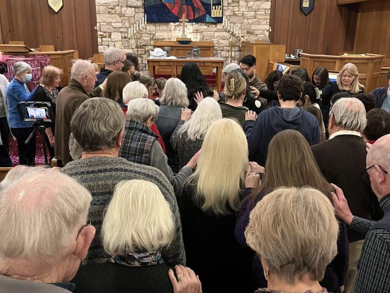 A group of people gather closely in a wooden room with stone decorations, engaged in what appears to be a communal or religious activity.