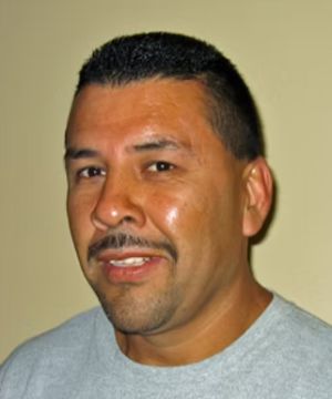 A person with short black hair and a mustache is smiling. The background is a plain beige wall. No landmarks are visible.