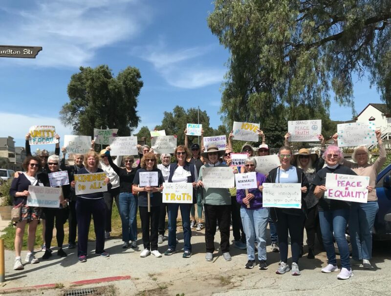 A group of people holding signs advocating for peace, truth, and equality on a sunny day near Arlington Street, with trees in the background.