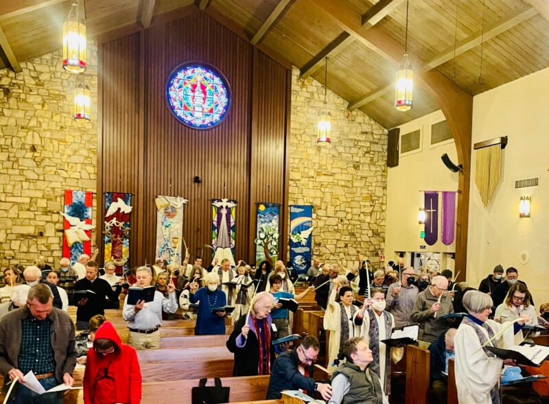 A church interior with a group of people holding palm leaves during a service. Stained glass and colorful banners adorn the walls.
