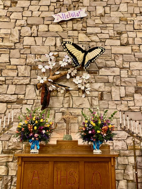 Stone wall with butterfly and flower decor, wooden altar below. "Alleluia" banner above, colorful floral arrangements on either side, and candlesticks present.