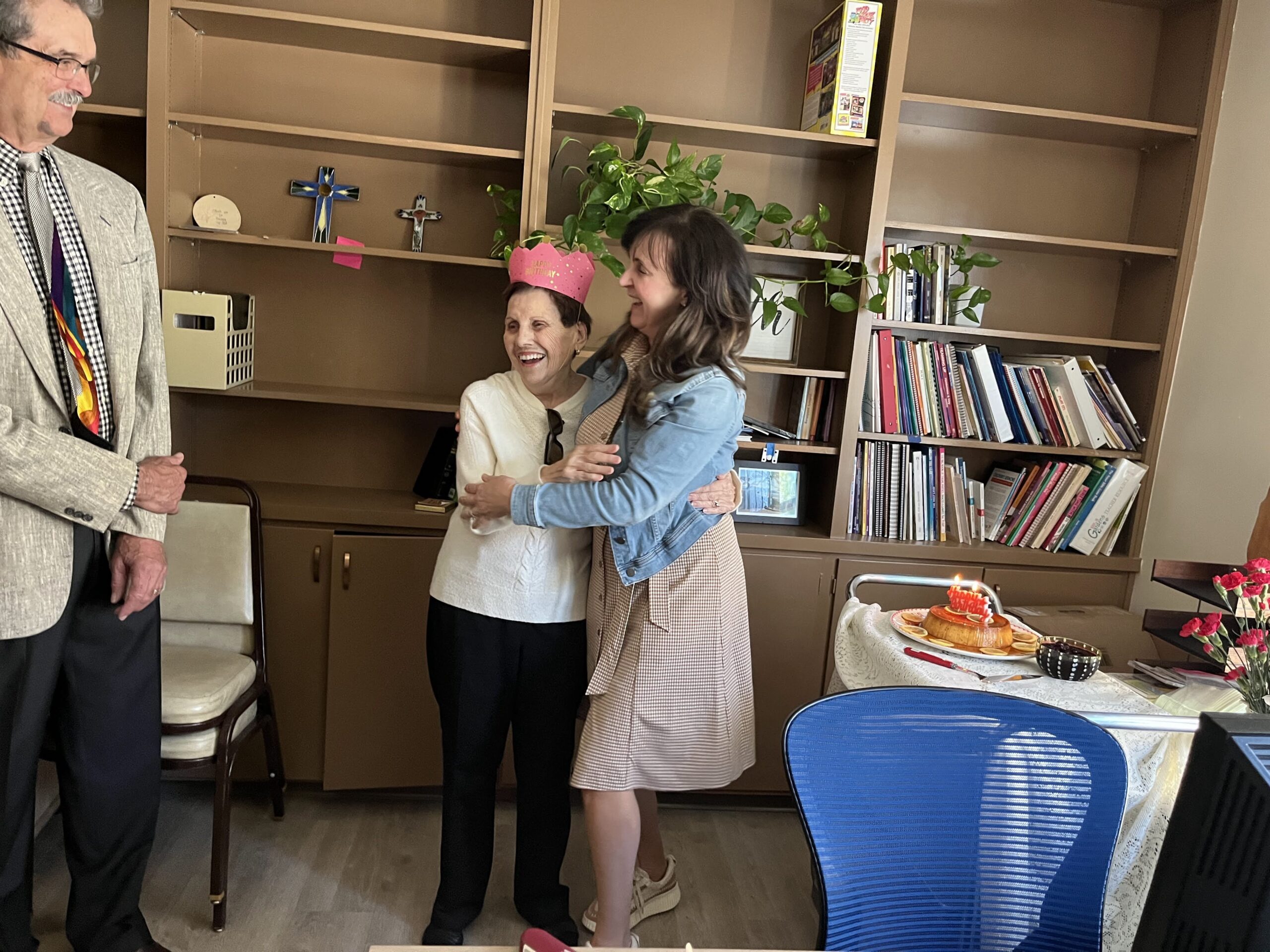 Three people enjoy a celebration in an office setting with bookshelves, decorations, and a small table with a cake and candles.