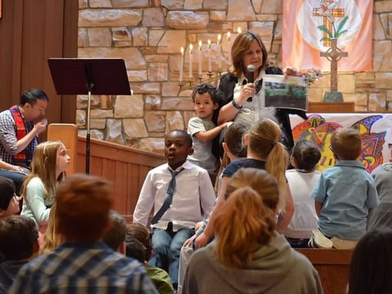 A person speaks to children in a church setting, holding a microphone and photo. Candles and colorful artwork in the background.