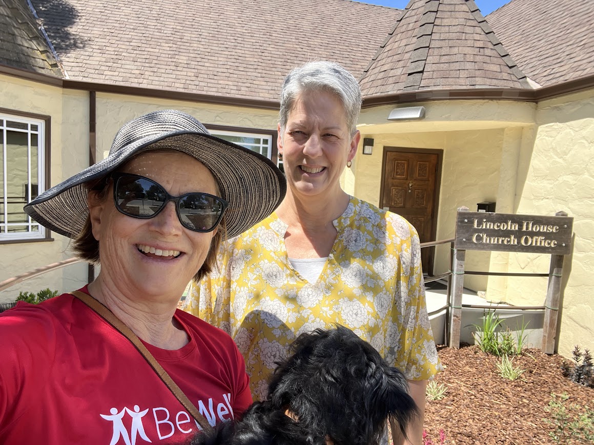 Two people and a dog in front of Lincoln House Church Office, smiling, with sunlight casting shadows on the building's textured wall.