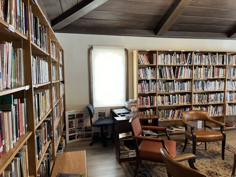 Cozy library interior with wooden bookshelves filled with books, two leather chairs, and a small desk near a sunlit window.