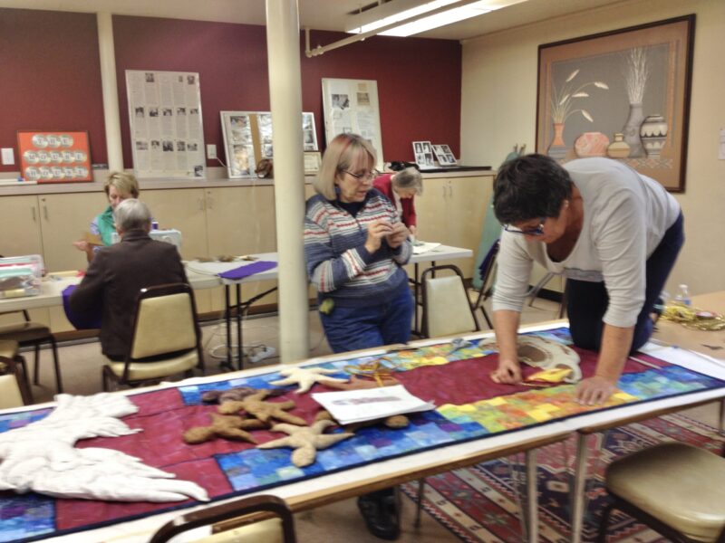 Several people engage in crafting activities at tables in a room adorned with large artwork and newspaper clippings on the walls.