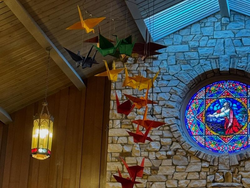 Stone interior features colorful origami cranes and a stained glass window depicting a biblical scene. A decorative hanging light is visible.