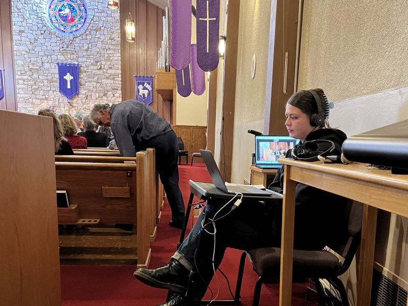 A person manages audio equipment in a church with wooden pews, stained glass, and religious banners. Another person leans over a pew.