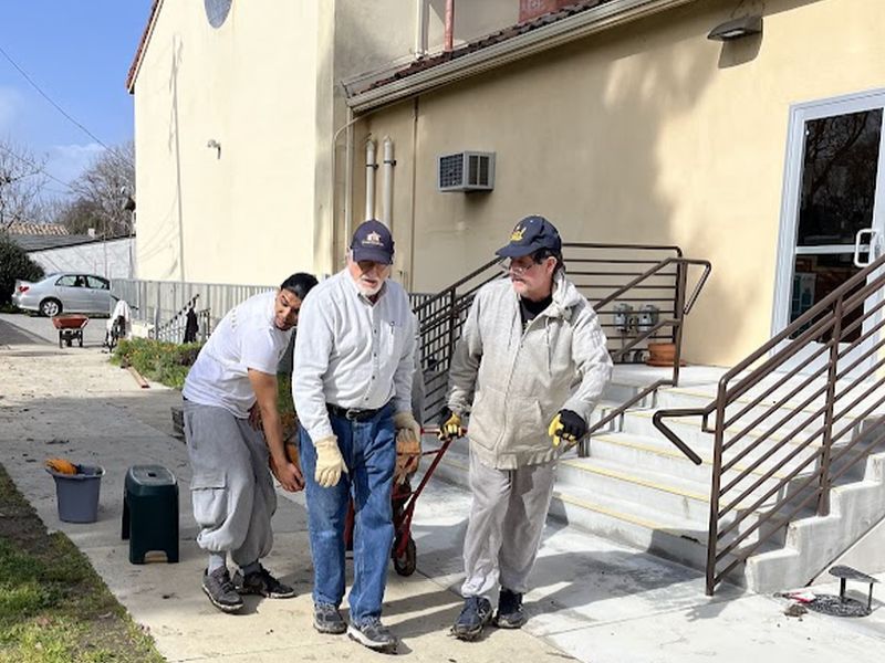 Three people work together, moving items outside a beige building with stairs. They wear casual clothing and appear involved in a manual task.