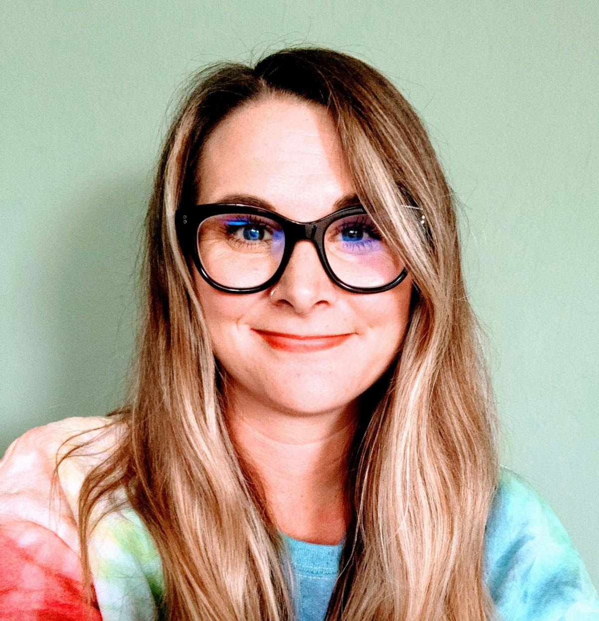 A person with long hair and glasses smiles against a plain green background, wearing a colorful tie-dye shirt.