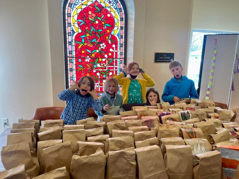 Five children making funny faces behind a table filled with decorated brown bags. A stained glass window adds color to the background.