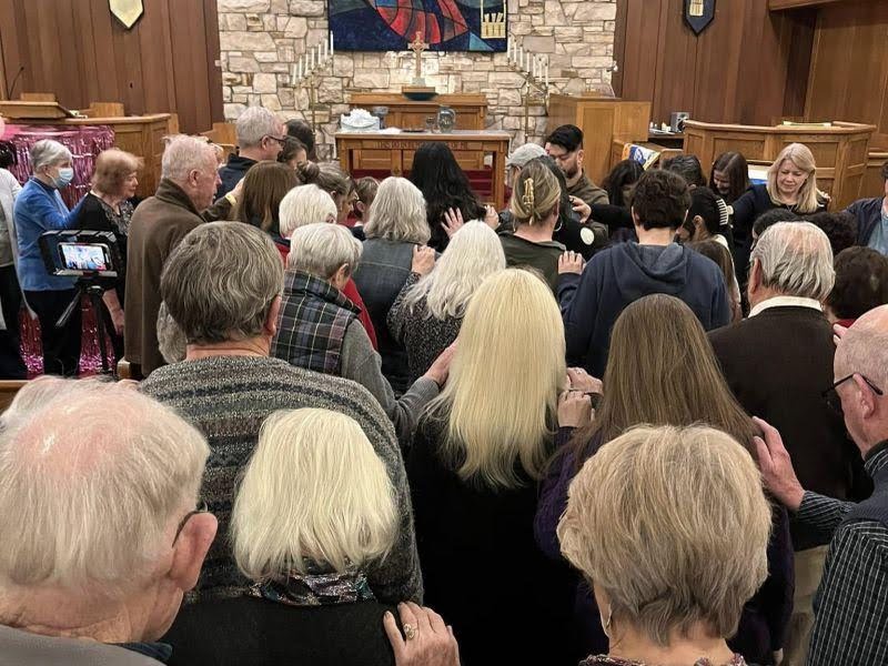 A group of people gathered in a church, standing close together, possibly in prayer or fellowship, with a stone wall in the background.