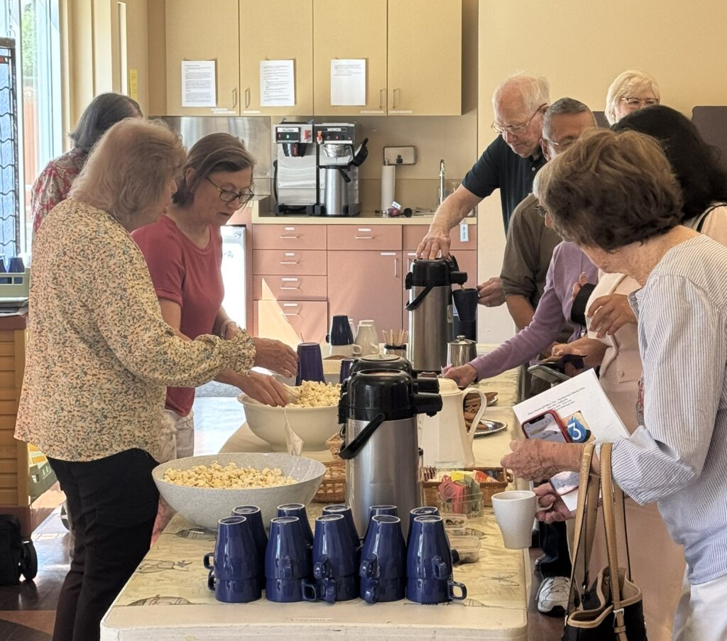 People gathering around a table with snacks and coffee, engaging in conversation. A clock is visible on the wall in the room.