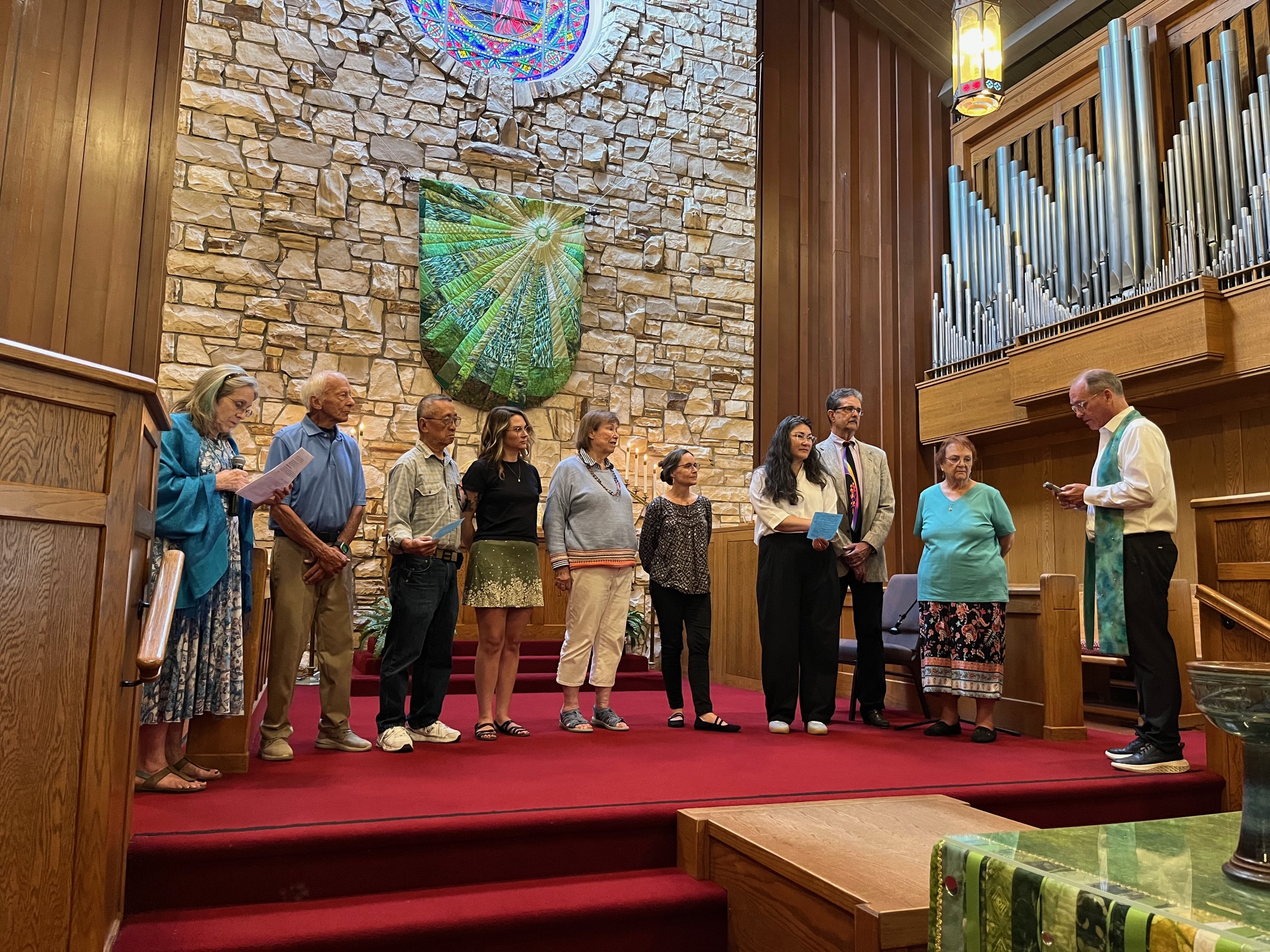 A group of people stand inside a church with stained glass, a stone wall, and organ pipes, possibly during a ceremony or service.