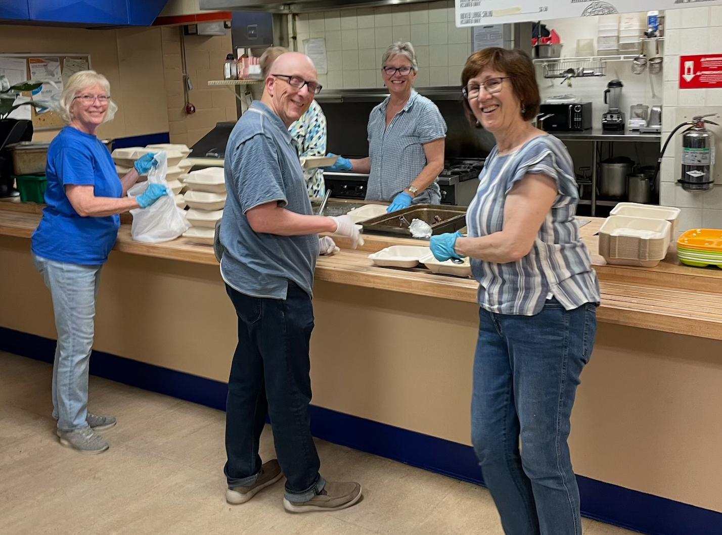 Four people smile while preparing meals in a kitchen, wearing gloves, surrounded by food containers on a wooden counter.