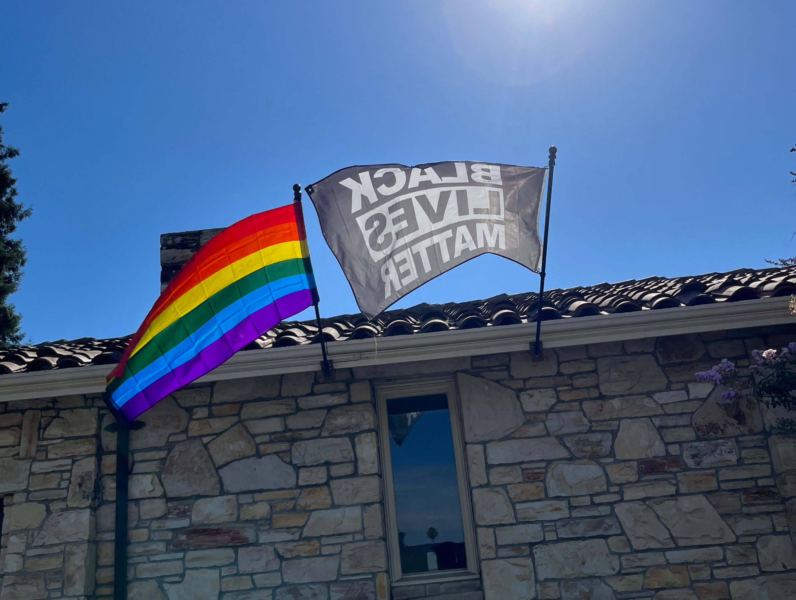 A stone house with Black Lives Matter and rainbow flags displayed under a clear blue sky, symbolizing support for equality and diversity.