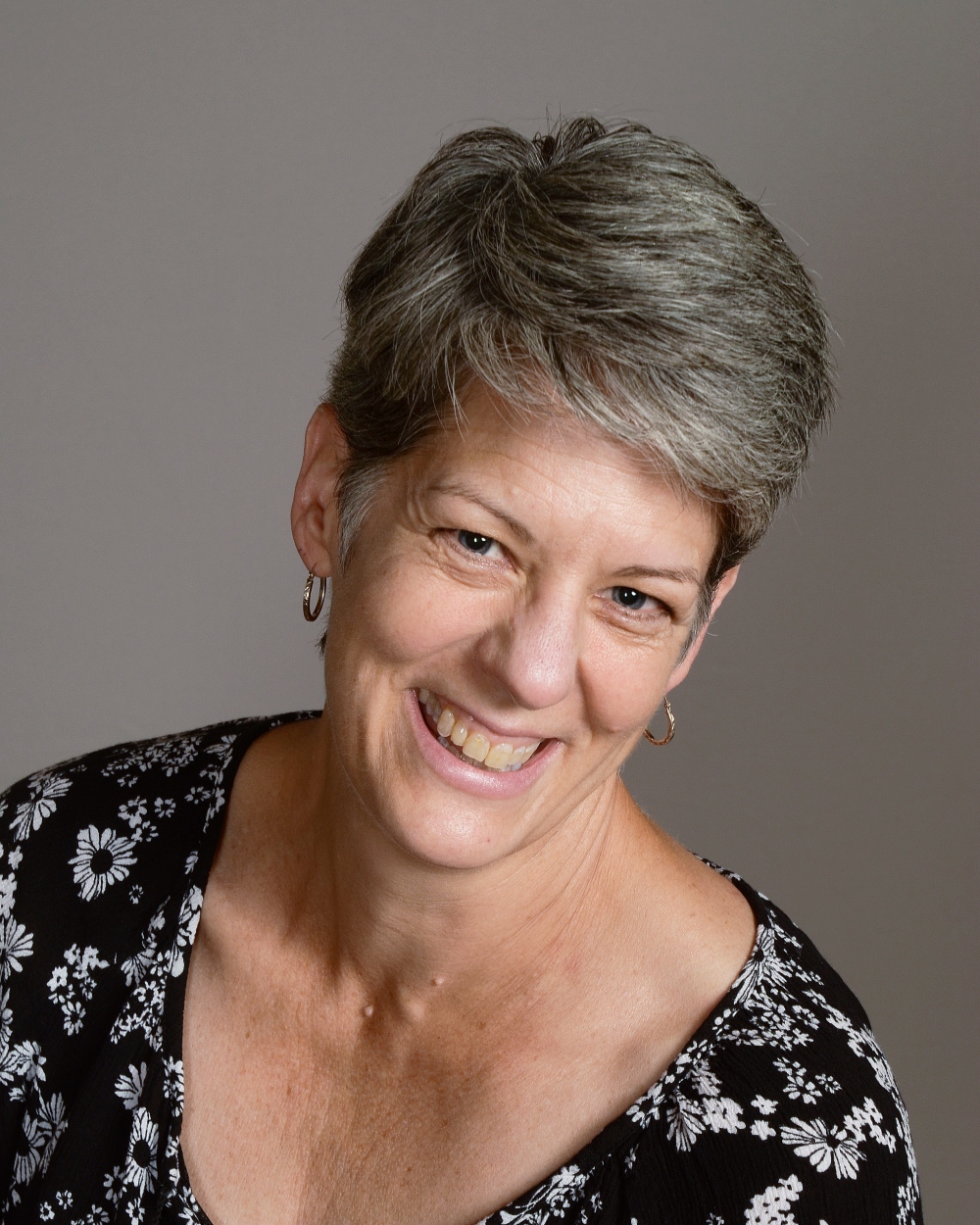 A smiling person with short, gray hair wearing a floral-patterned shirt poses against a plain background, exuding warmth and friendliness.