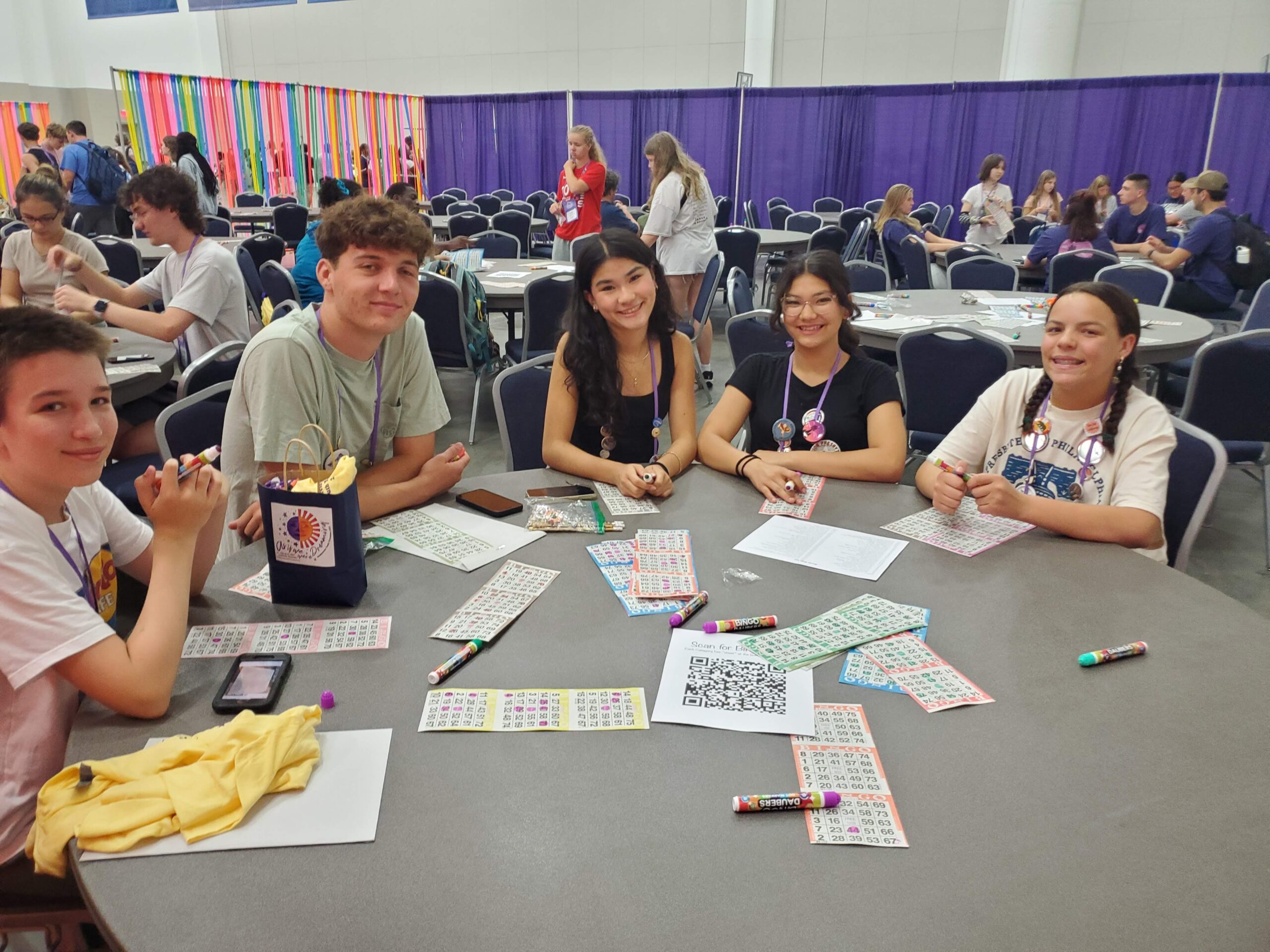 A group of people sitting at a table with bingo cards and daubers, in a convention hall setting with colorful decorations.