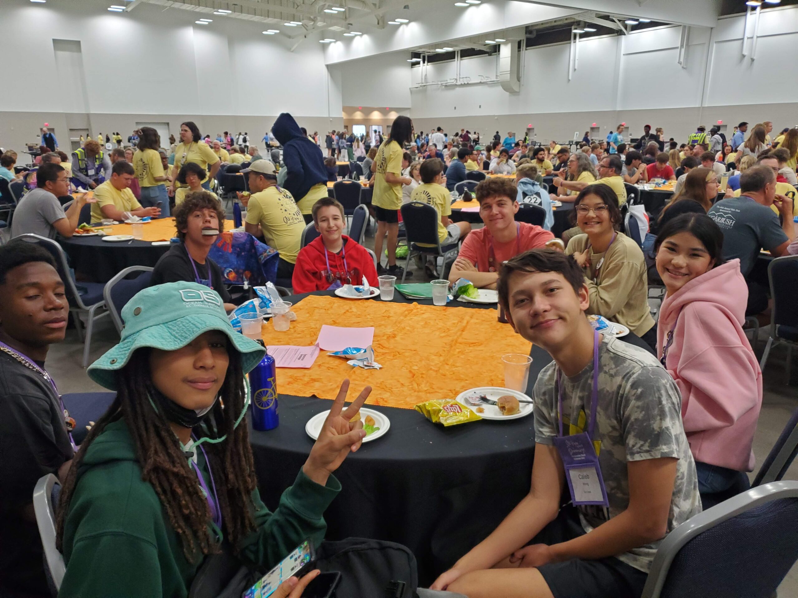 A large gathering of people in a spacious hall, sitting at round tables, enjoying food and conversation. Vibrant and social atmosphere.