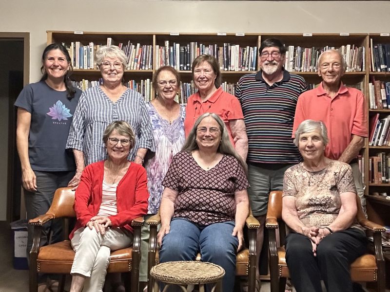 Nine people posing in a library, with bookshelves in the background. Some are seated, others standing, all smiling at the camera.