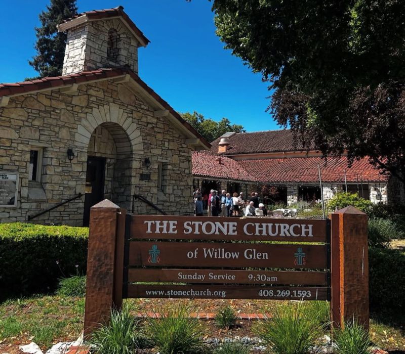 A group of people gather outside The Stone Church of Willow Glen, a stone building with a prominent entrance under a clear sky.